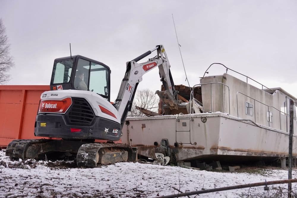 Bobcat excavator removing debris from a rundown boat in a snowy environment.