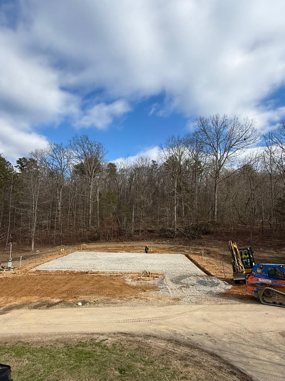 Construction site with gravel foundation surrounded by trees under a cloudy sky.
