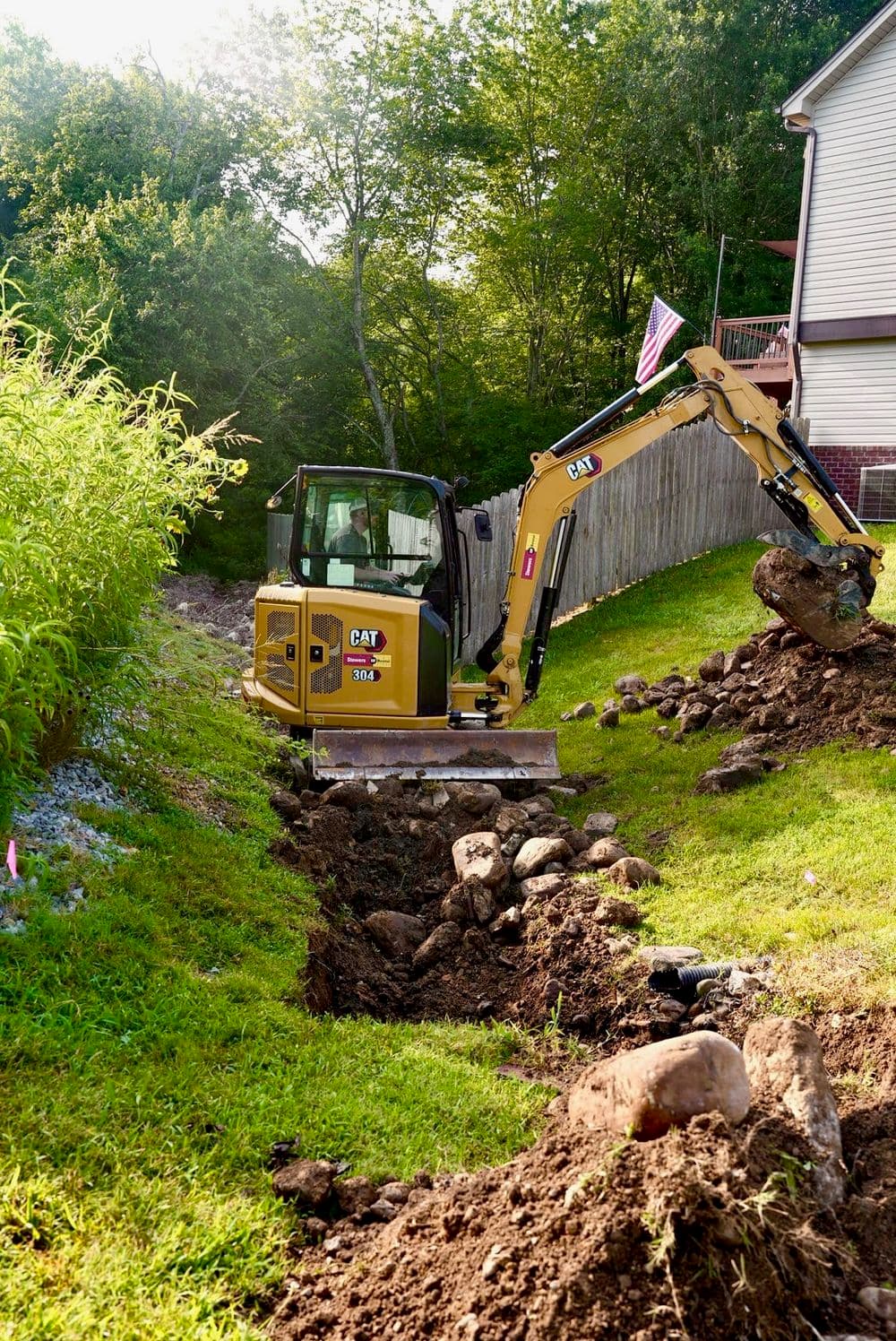 Excavator digging a trench in a residential yard with rocks and greenery nearby.