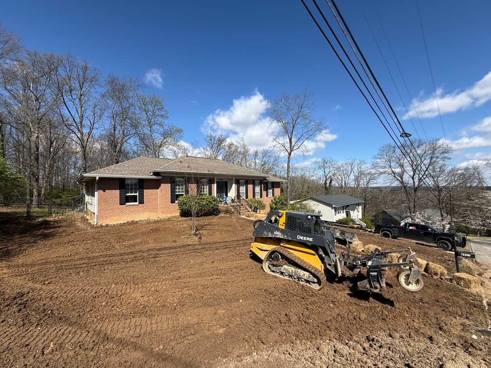Excavator near a house on a hillside, clearing land with trees in the background.