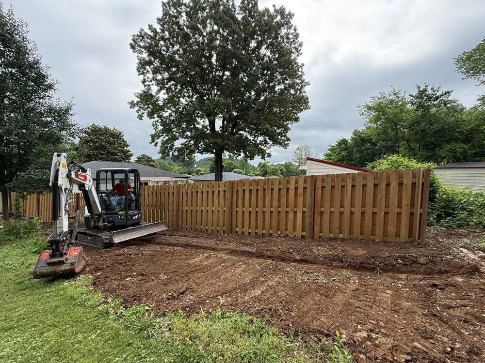 Excavator leveling soil near wooden fence in residential backyard.