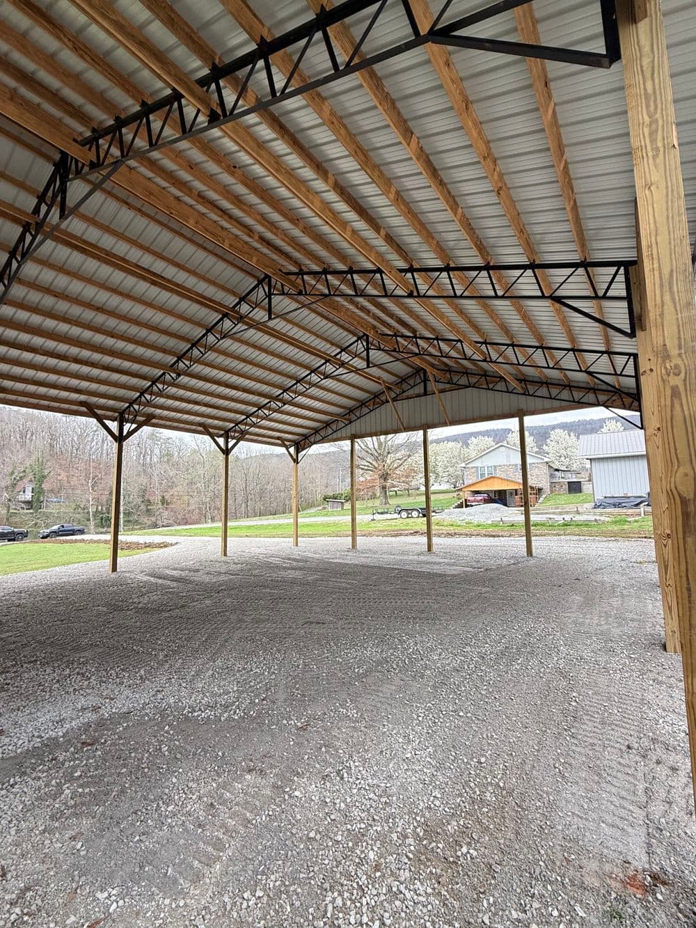 Open metal pole barn interior with gravel floor and wooden support beams in a rural setting.