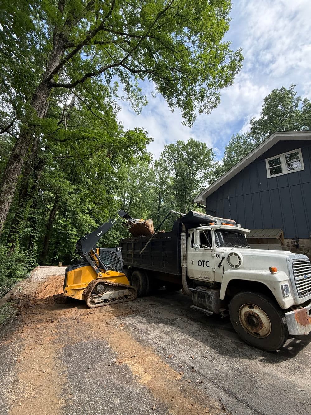 Dump truck unloading dirt near a construction site surrounded by trees and a building.