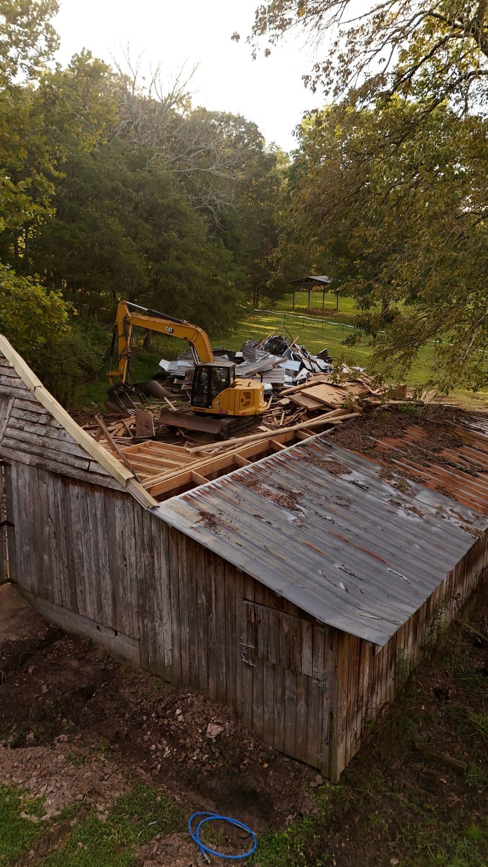 Excavator demolishing a wooden barn with debris in a lush green landscape.