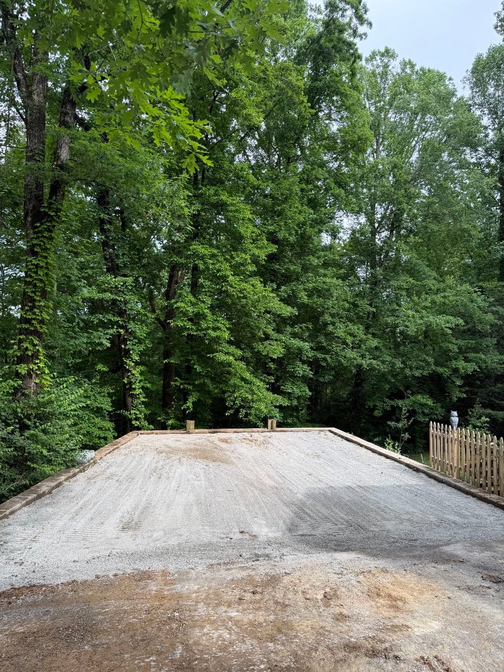 Gravel driveway preparation in a wooded area, surrounded by lush greenery.