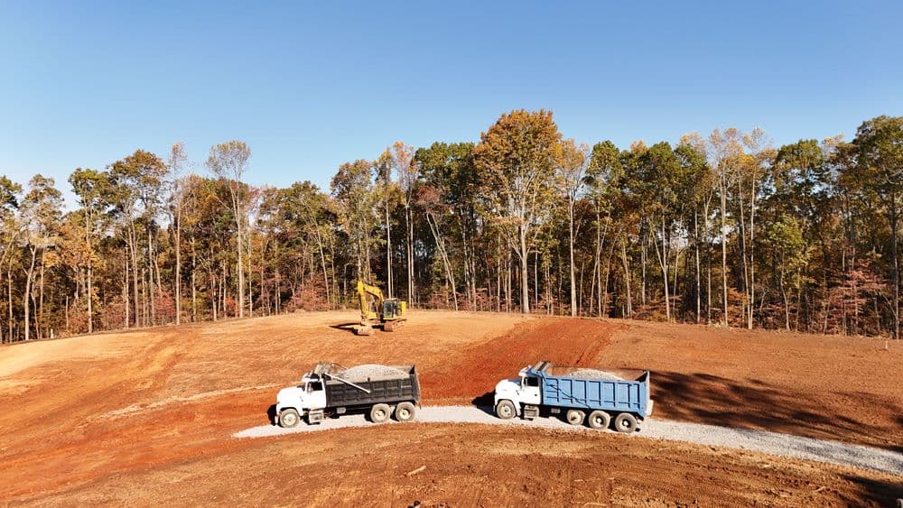 Construction site with dump trucks and excavator on a gravel road amidst autumn trees.