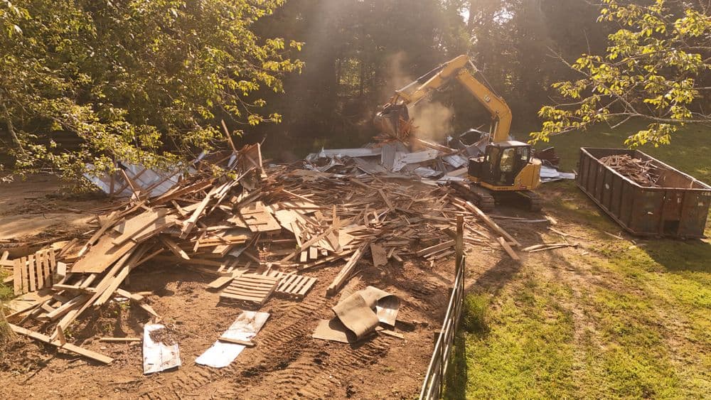 Excavator demolishing wooden structure, debris scattered, construction site in background.